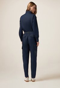 Jumpsuit in denim con colletto, vita elastica e tasche cargo. Colore blu scuro con accenti di strass sulle tasche laterali.