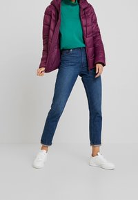 Lekkie, pikowane fioletowe kurtka puchowa z kapturem noszona na zielonym golfie, w połączeniu z wysoko stanowanymi niebieskimi dżinsami i białymi sneakersami.