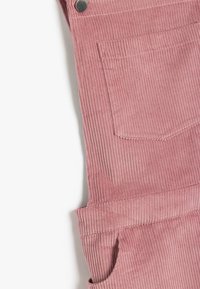 Rosa corduroy-overaller med vertikalt ribbad textur, sidofickor och metallknappar. Tyget ser mjukt och slitstarkt ut.