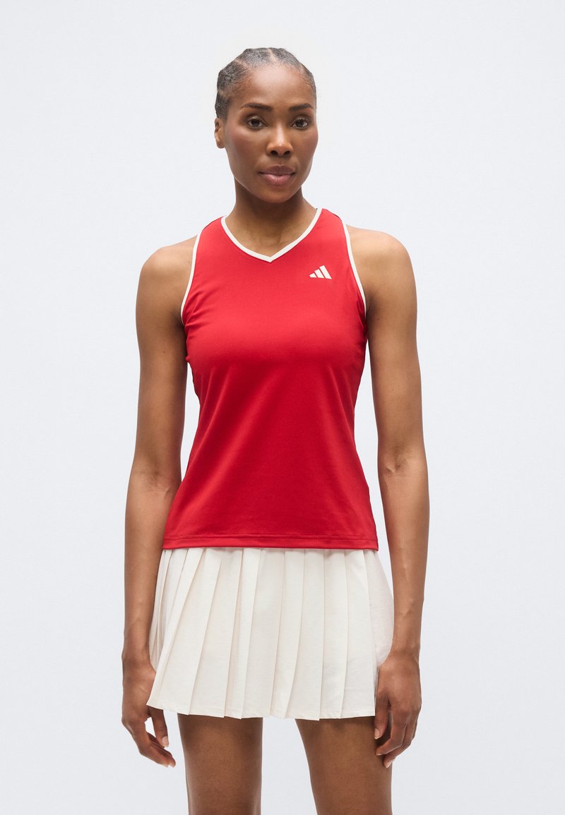 Haut de sport rouge sans manches avec bordure blanche et logo latéral, associé à une jupe plissée crème. Le tissu semble lisse et léger.