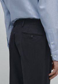 Navy broek met een gladde textuur, met een achterzak met een knoop. Gecombineerd met een lichtblauw gestreept overhemd.