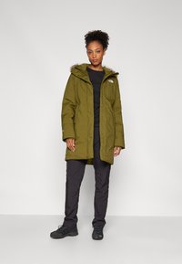 Olivově zelená parka s kapucí lemovanou kožešinou, zapínání na zip a knoflíky a bočními kapsami, nošena přes černý top a černé kalhoty.