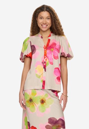 Kobieta o kręconych włosach w beżowym zestawie bluzki i spódnicy z dużymi, kolorowymi kwiatowymi wzorami, uśmiechająca się, z rękami swobodnie opuszczonymi wzdłuż ciała.
