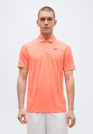 Modèle masculin portant un polo Nike corail à manches courtes avec un logo swoosh noir, associé à un short blanc, debout devant un fond clair uni.