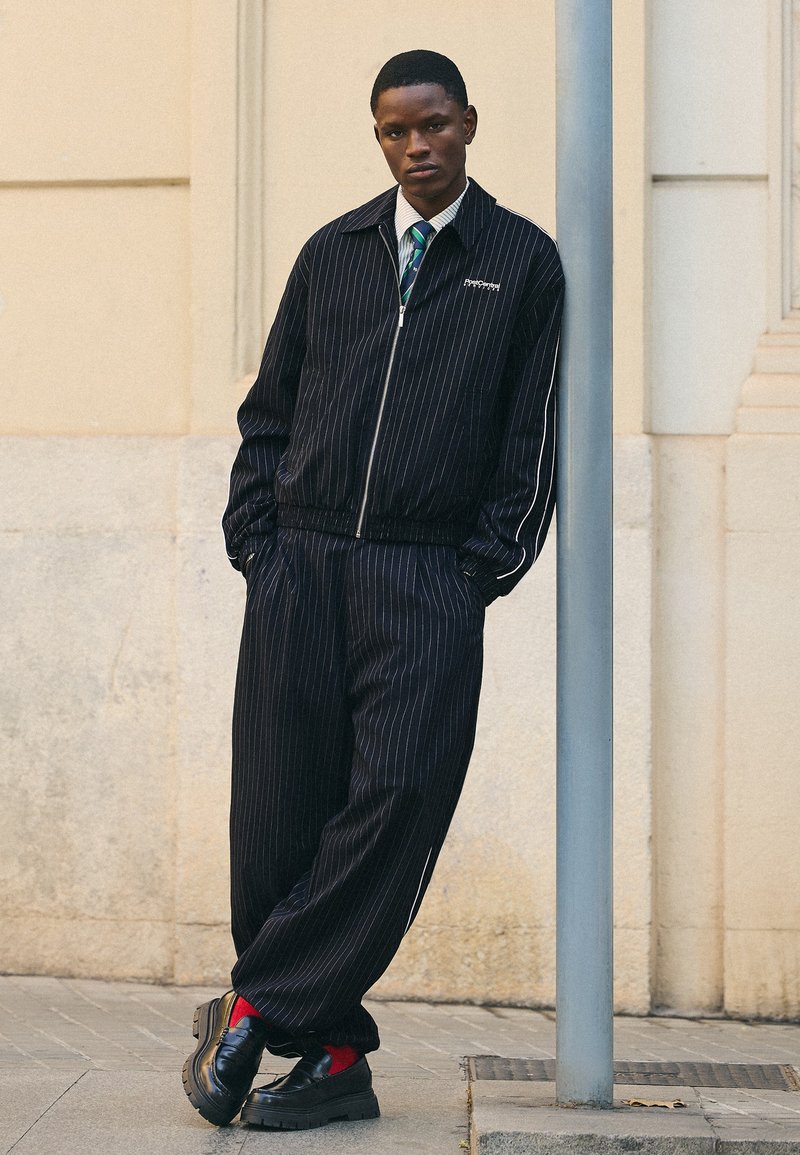 Jeune homme appuyé contre un poteau, portant une veste et un pantalon noirs à fines rayures, une chemise blanche, une cravate rayée, des chaussettes rouges et des chaussures noires.