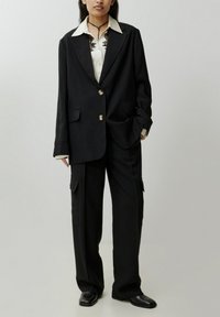 Zwarte tailored blazer met gouden knopen, witte shirt met zwarte borduursels, en losse zwarte cargobroek. Glad, formeel stof.