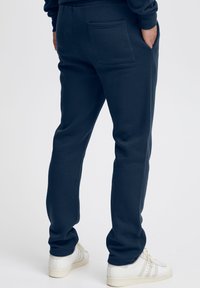 Marineblaue Baumwoll-Jogginghosen mit lockerem Schnitt, ausgestattet mit einem elastischen Bund, Seitentaschen und einer einzelnen Gesäßtasche. Weiße Turnschuhe.