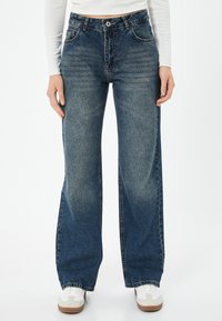 Siniset korkeat farkut leveällä lahkeella, joissa on vaalea kuluminen, viisi taskua ja sävyttävä ommel, yhdistettynä valkoisiin lenkkareihin.