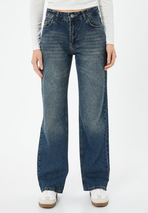 Blauwe high-waisted jeans met een wijde pasvorm, voorzien van lichte vervaging, vijf zakken en ton-sur-ton stiksel, gecombineerd met witte sneakers.
