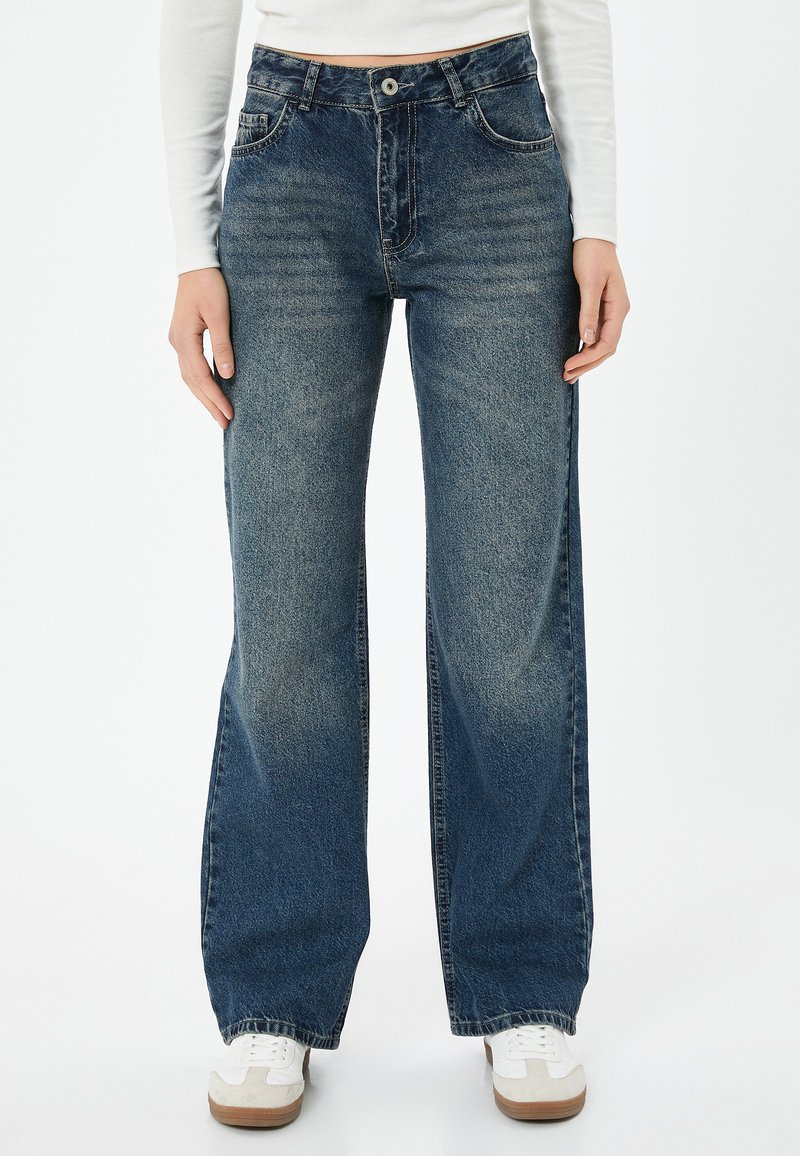 Siniset korkeat farkut leveällä lahkeella, joissa on vaalea kuluminen, viisi taskua ja sävyttävä ommel, yhdistettynä valkoisiin lenkkareihin.