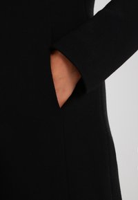 Vestido de tela negra con un bolsillo lateral, que presenta una textura suave y un diseño de mangas ajustadas. La tela tiene un ligero estiramiento para mayor comodidad.
