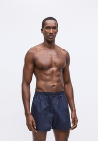 Muskuløs mann som står mot en ensfarget bakgrunn iført marineblå boxer-shorts med svart midjebånd.