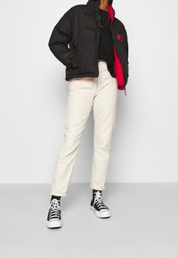 Sort quiltet pufferjakke med rød indvendig foring, sort t-shirt, beige lige ben jeans og sorte højtop sneakers med hvide såler.