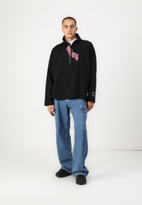 Black pullover with a high collar and front floral print, paired with loose blue jeans and black shoes. Simple design, relaxed fit.
