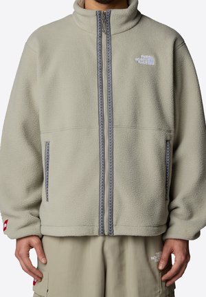 Veste polaire beige avec fermeture éclair avant, col haut, deux poches zippées, et logo The North Face sur la poitrine et le pantalon.