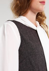 Chaleco oscuro texturizado con una blusa blanca clara debajo, que presenta un cuello atado y una vista en primer plano de los detalles de la tela y el escote.