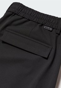 Detailní záběr na černé kalhoty s elastickým pasem a klopovou kapsou, na které je malá obdélníková visačka s logem "MNG".