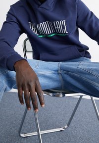 Mörkblå hoodie med "The North Face"-logotyp, kombinerad med ljusblå denimjeans, sittande på en metallstol med kromade detaljer.