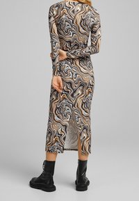 Žena v šatech s dlouhým rukávem do délky lýtek, které mají hnědý, černý a bílý vícestarý vzor, v kombinaci s černými botami na šněrování.