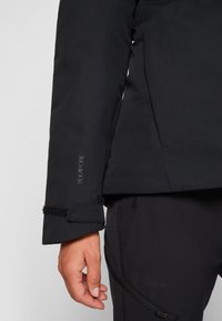 Gros plan sur le bras gauche d’une personne portant une veste Texapore noire avec une bride de poignet réglable, main détendue le long du corps, associée à un pantalon noir.