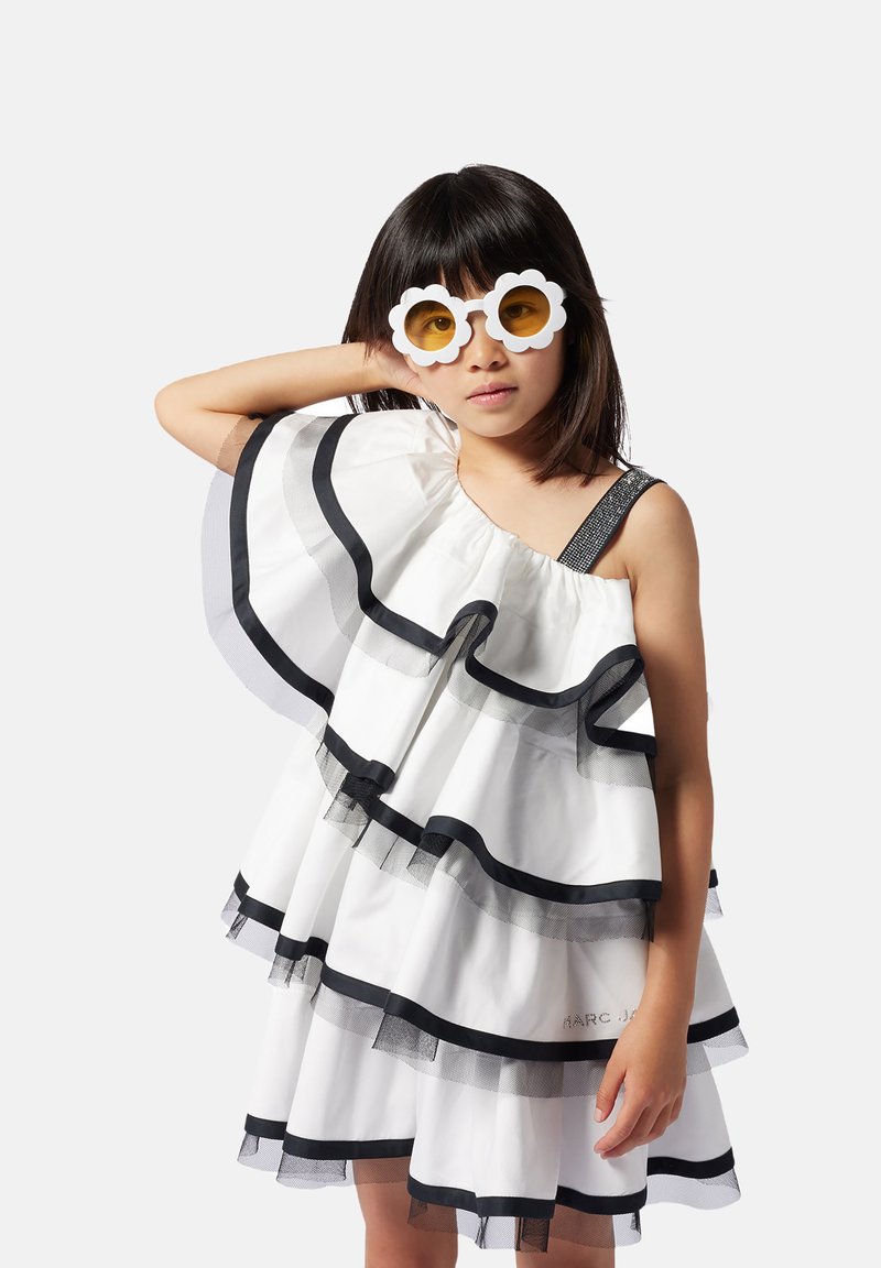 Robe blanche avec des volants noirs et des accents en tulle, avec une seule bretelle, ornée d'une épaule à sequins. Complétée par des lunettes de soleil en forme de fleur.