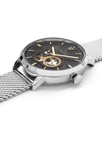 Montre avec bracelet en mailles argentées, cadran noir, accents dorés, design squelettique et engrenages visibles, avec une couronne sur le côté.