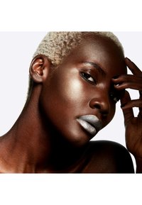 Gros plan sur une personne à la peau foncée, avec des cheveux blonds texturés et courts, du rouge à lèvres argenté et un highlighter lumineux sur les joues et la clavicule.