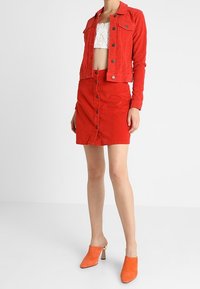 Giacca di velluto a coste rossa con bottoni argentati, gonna aderente in velluto a coste rossa con chiusura anteriore a bottoni, e mule arancioni con tacco. Sotto, un top corto in pizzo bianco.