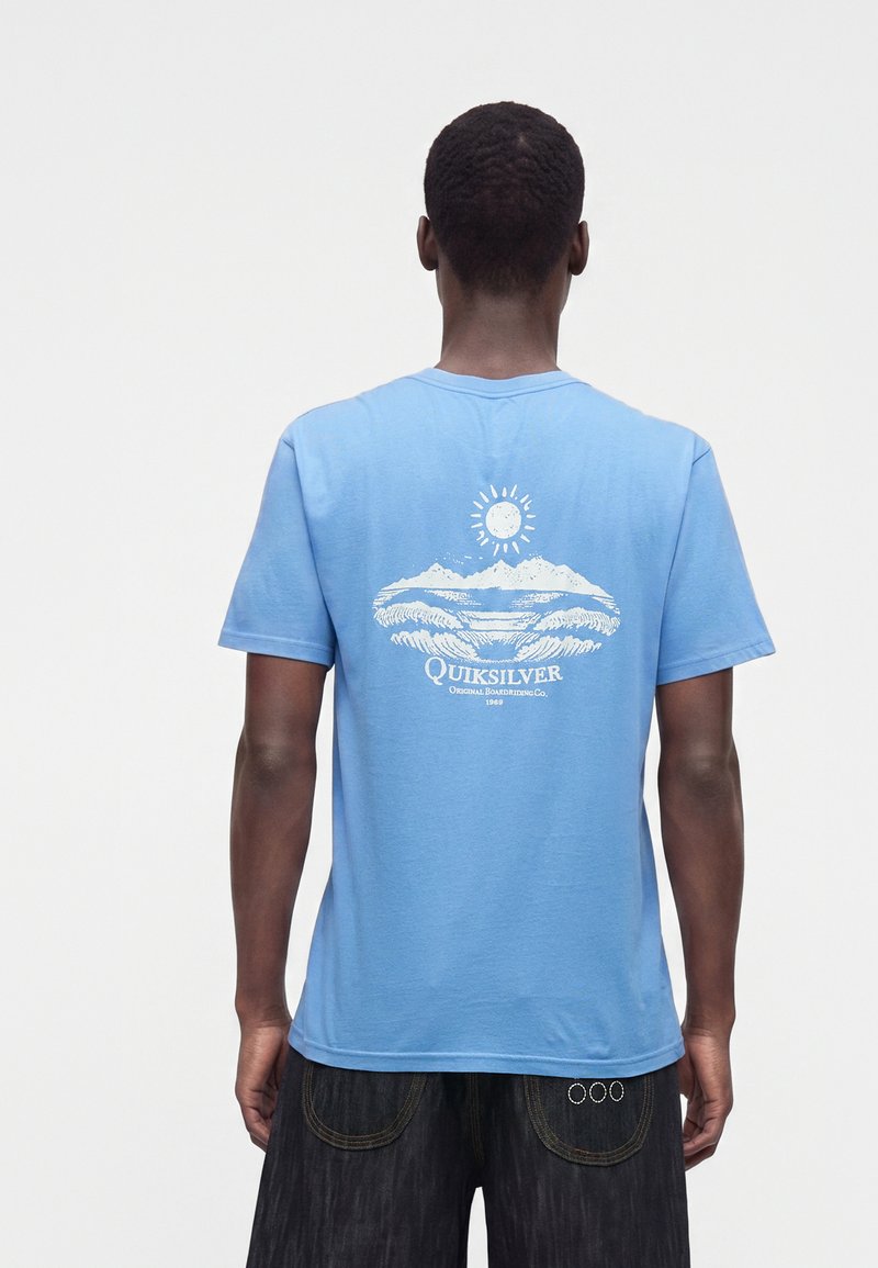 Άνδρας που φορά ένα γαλάζιο t-shirt Quiksilver με λευκό σχέδιο ήλιου και κύματος στην πλάτη, συνδυασμένο με σκούρα σορτς, γυρισμένος με την πλάτη.