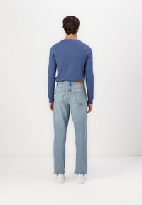 Un pull bleu à manches longues associé à un jean clair et des baskets blanches, vu de dos. Présente un tissu texturé et un patch en cuir marron.