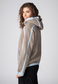 Fleecevest in beige met lichtblauwe accenten. Voorzien van een capuchon, een met rits afsluitbare zijzak en een gestructureerde stof. Ruime pasvorm, licht oversized ontwerp.
