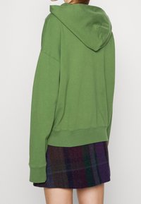 Groene katoenen hoodie met een capuchon, ontspannen pasvorm en geribde manchetten. Draagt over een donkere geruite rok met paarse en groene tinten.