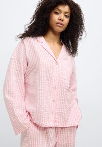 Σετ ροζ πιτζάμες με καρό σχέδιο που περιλαμβάνει πουκάμισο με κουμπιά και τσέπη, με άνετη εφαρμογή. Το ύφασμα φαίνεται υφασμένο με ελαφριά, αεριζόμενη ποιότητα.