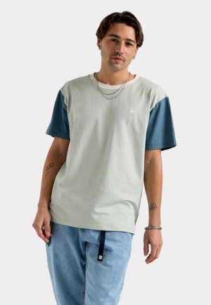 Joven con cabello corto y oscuro que lleva una camiseta de dos tonos en gris y azul, jeans azul claro, cadenas plateadas y muestra tatuajes florales en ambos antebrazos.