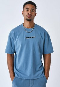 Camiseta de algodón azul con mangas cortas, escote redondo y un logotipo "PXP" en un parche rectangular en el centro. Textura suave.