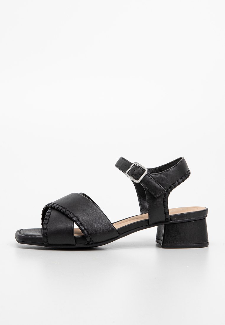 Sandalia de cuero negra con detalles trenzados, punta abierta, correa ajustable en el tobillo y un tacón bajo. Textura suave y diseño minimalista.