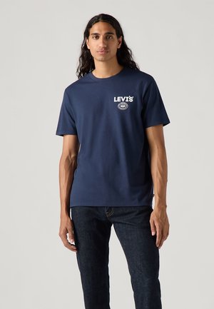 Tamsiai mėlynas medvilninis marškinėliai trumpomis rankovėmis, apvaliu kaklu ir baltu Levi's logotipu ant krūtinės. Dėvima kartu su tamsiomis džinsomis.