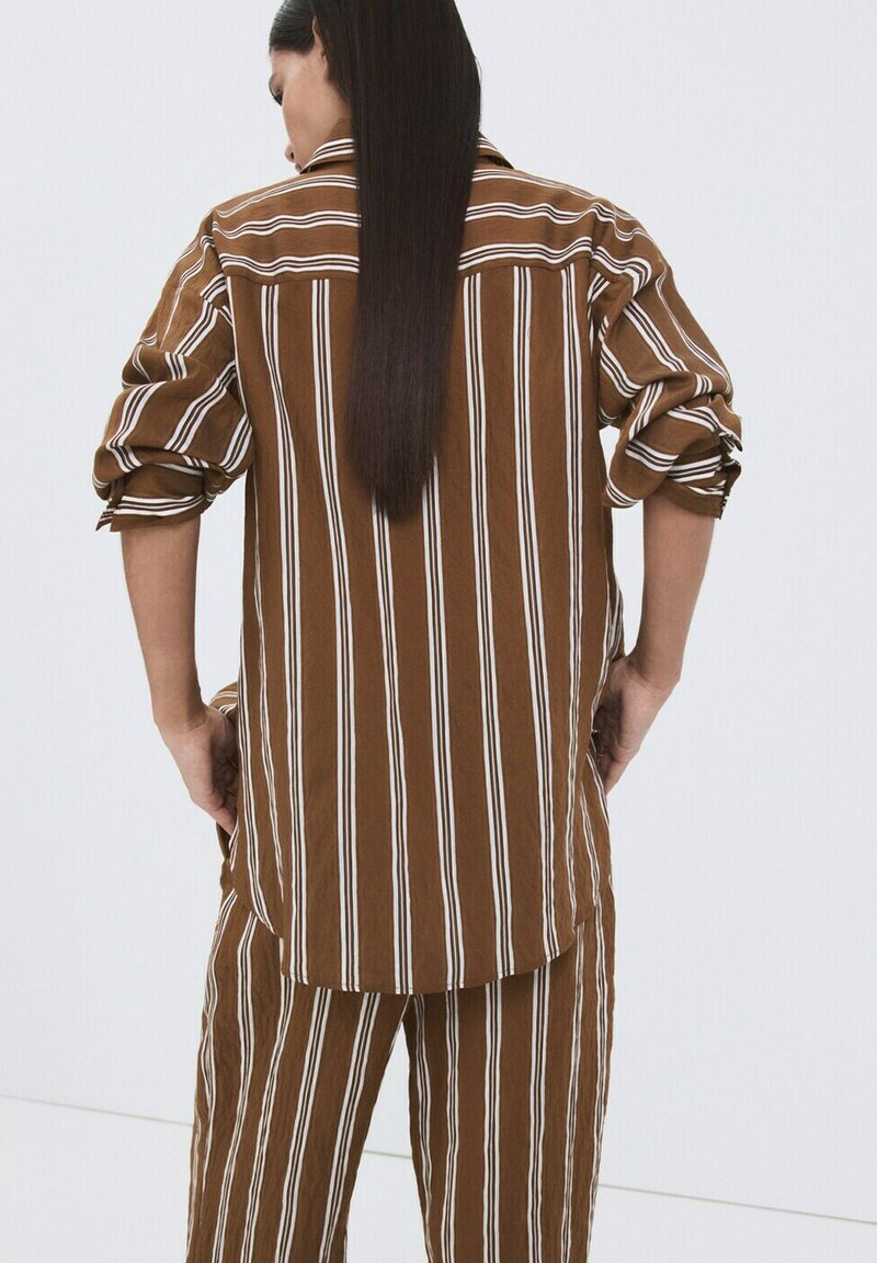 Mujer con cabello largo y liso, vistiendo camisa y pantalones con rayas verticales marrones y blancas, manos apoyadas en las caderas, de espaldas.