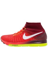 Červené pletené tenisky s vysokým designem kotníku, mají bílé logo Nike Swoosh, žluté tkaničky a zeleně zabarvenou podrážku s označením "Nike Zoom".