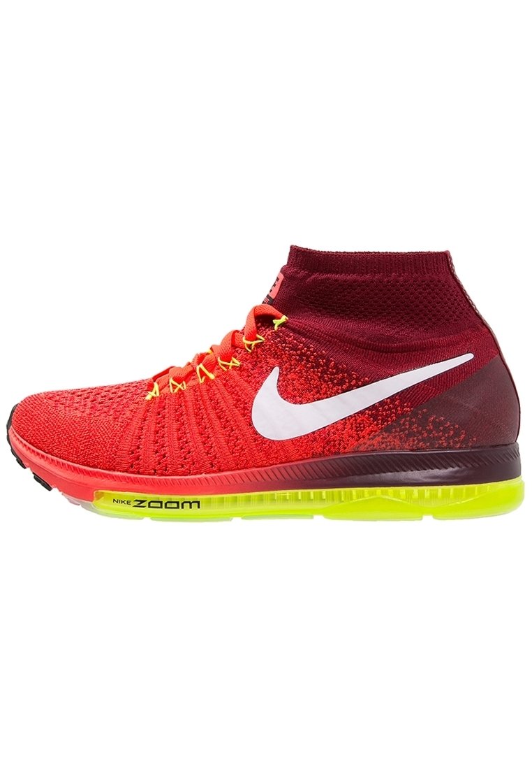Červené pletené tenisky s vysokým designem kotníku, mají bílé logo Nike Swoosh, žluté tkaničky a zeleně zabarvenou podrážku s označením "Nike Zoom".