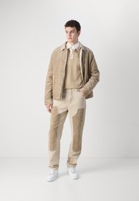 Béžová kordová bunda, zodpovedajúci béžový sveter a nohavice s semišovými nášivkami. Biely športový obuv dopĺňa outfit na svetlom pozadí.
