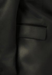 Schwarze Jacke aus glattem Stoff, mit einer einzelnen Brusttasche, subtil strukturierter Oberfläche und taillierter Passform.
