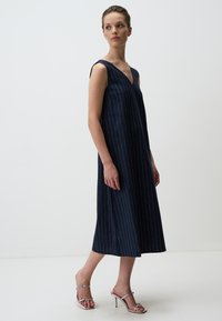Donkerblauwe mouwloze midijurk met verticale witte strepen, diepe V-hals, losse pasvorm, zachte stof, gecombineerd met zilveren hoge hakken.