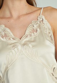 Camisón de satén color crema con un intrincado detalle de encaje floral en el escote y el pecho, que lleva una persona con los hombros al descubierto.