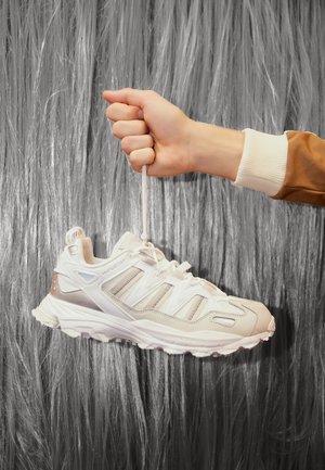 Ręka trzyma beżowego sneakersa za sznurówki na teksturowanym szarym, pionowym tle.