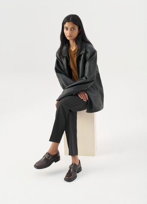 Mujer con cabello largo y negro sentada sobre un bloque claro, vistiendo una chaqueta de cuero negra, top marrón, pantalones negros y zapatos marrón oscuro con hebilla.