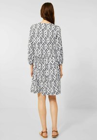 Robe blanche avec un motif géométrique bleu marine, manches longues, taille élastique et jupe évasée. Portée avec des sandales plates marron.