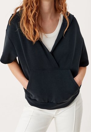 Kobieta ubrana w czarną bluzę z krótkim rękawem i kapturem z przednią kieszenią, na białym prążkowanym topie i białych spodniach, ręce w kieszeniach bluzy.