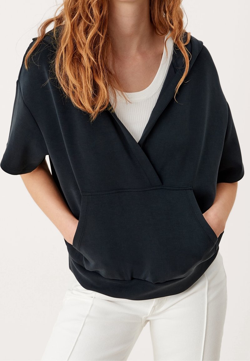 Femme portant un sweat à capuche noir à manches courtes avec une poche devant, sur un débardeur blanc côtelé et un pantalon blanc, les mains dans les poches du sweat.