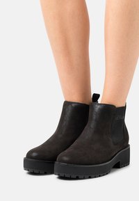 Schwarze Wildleder-Ankle-Boots mit einem klobigen Absatz, elastischen Seiteneinsätzen und einem Logo-Detail an der Außenseite. Strukturiertes Profil für besseren Halt.